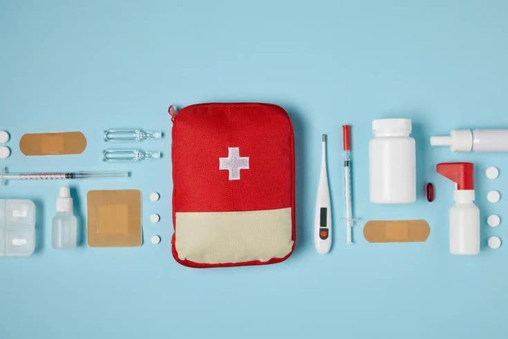 : Un botiquín de primeros auxilios rojo con un símbolo de cruz blanca en el centro, rodeado de varios suministros médicos organizados sobre un fondo azul. Entre los artículos hay vendas adhesivas, jeringas, frascos de medicamento, un termómetro, tabletas y ampollas de vidrio.